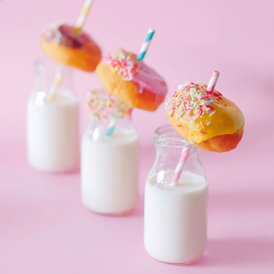Three small bottles of milk with small colourful donuts with sprinkles being held by colourful striped straws.