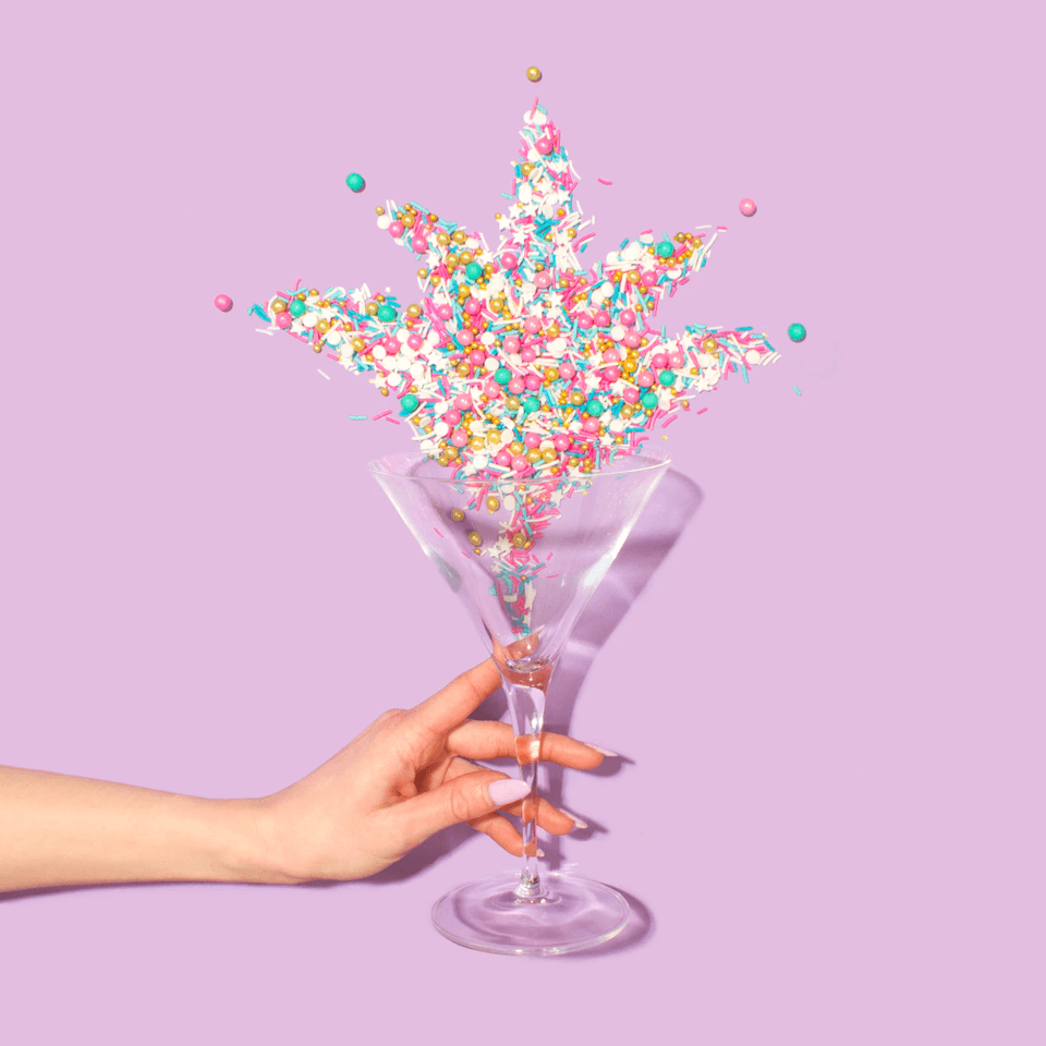 Lots of colourful sprinkles being thrown in the air from a champagne glass being held by a human with lilac nail polish.